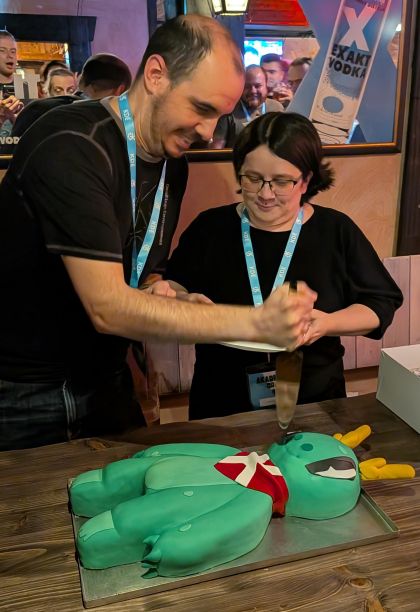 KDE e.V. president and vice-president cutting a cake in the shape of KDE's cute mascot Konqi with a large knife while visibly enjoying what they are doing.